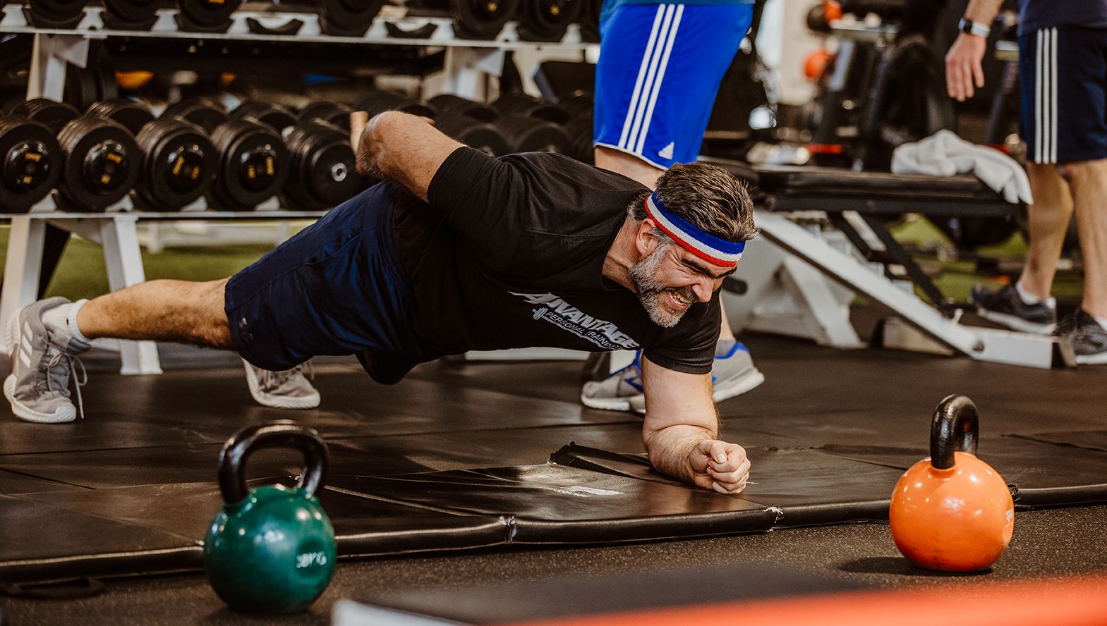 A member in an Advantage T-shirt and headband holds a plank beside two kettlebells