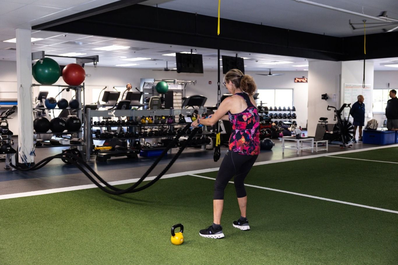 A member works the battle ropes on the turf, a kettlebell at her feet.
