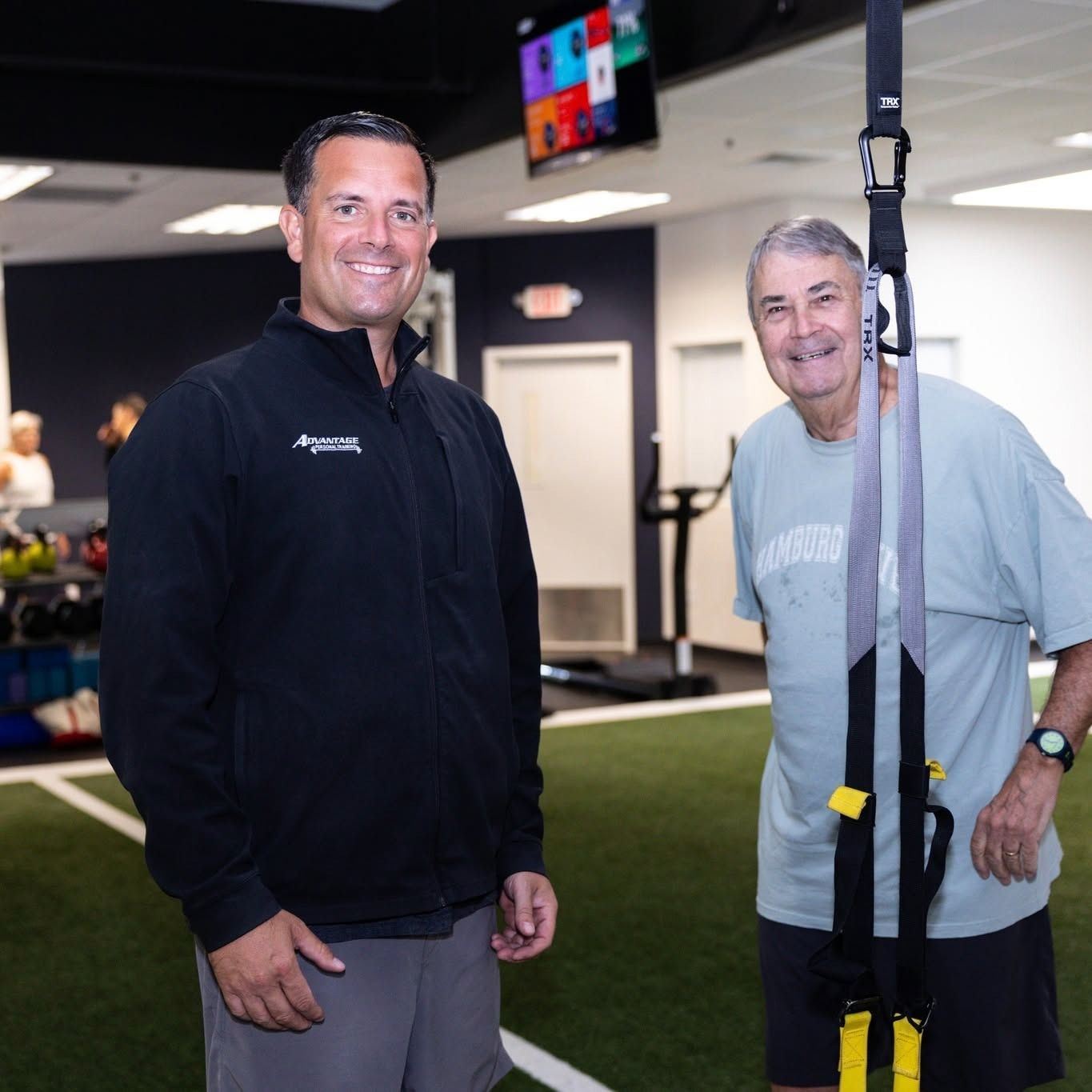 A smiling man in a black Advantage jacket stands beside an older member holding a suspension-trainer strap.