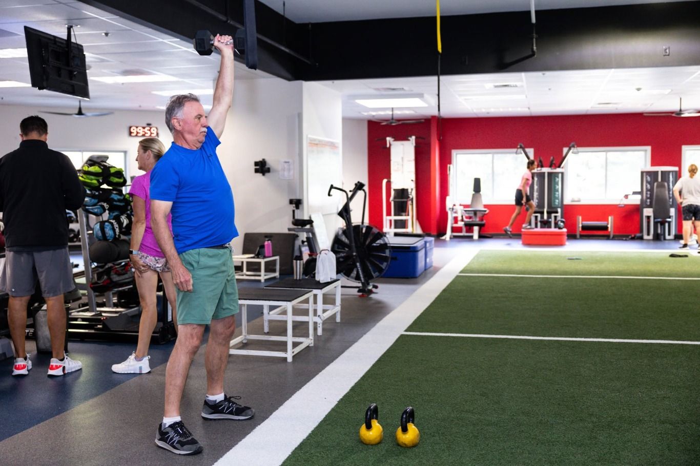 An older member presses a dumbbell overhead beside the turf while others train nearby; a red wall in the background.