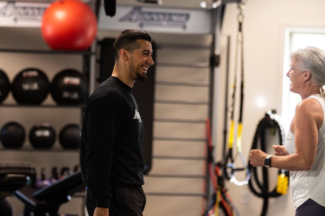 A coach and a member laughing together during a session by the medicine-ball rack.
