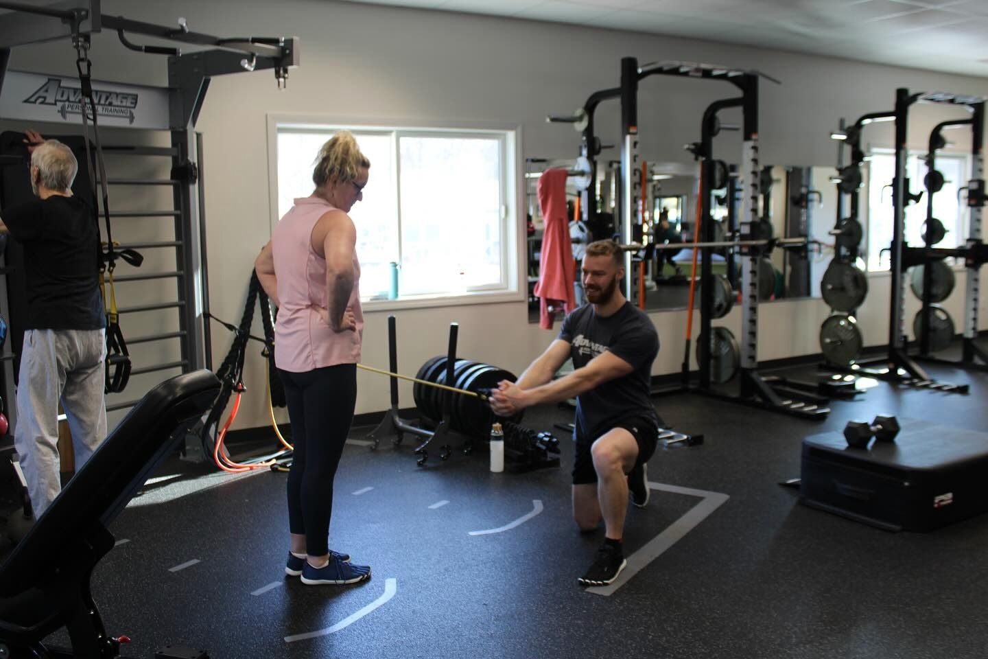 A coach in a half-kneeling stance holds out a resistance band to show a member the exercise; an older member trains at the wall bars behind them.